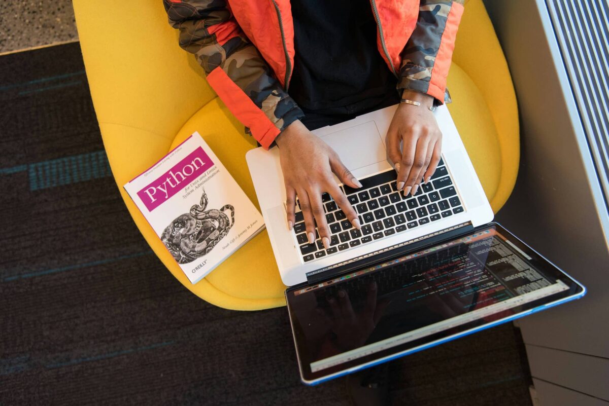 Imagen de una mujer escribiendo código en un ordenador con un libro de Python sobre la mesa.